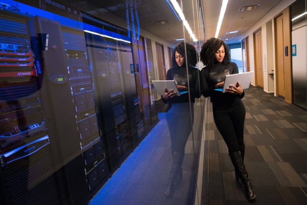 woman in black outfit holding laptop against a clear glass in a hallway with servers on the other side of the glass