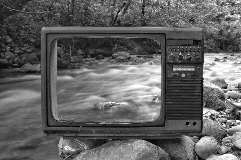 Black and white vintage old broken TV placed on stones near wild river flowing through forest